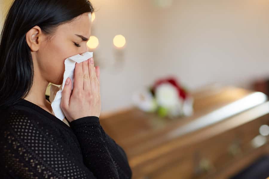 Millennial crying with tissue to her face and brown wooden casket behind her. Representing consequence of opioid crisis among millennials